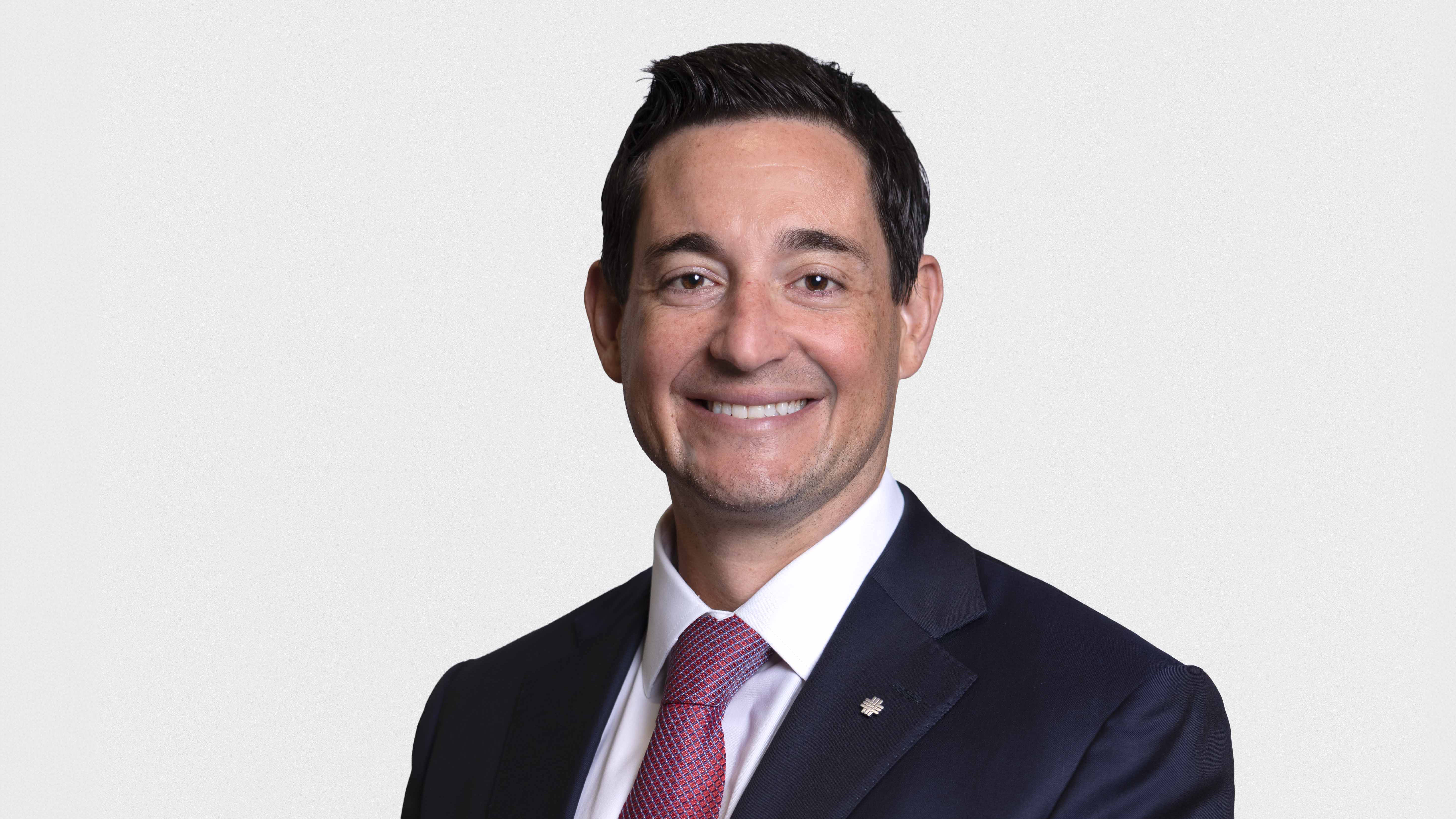 Studio portrait of an adult in a dark suit, white shirt, and red tie, standing against a plain light background with a small lapel pin visible.